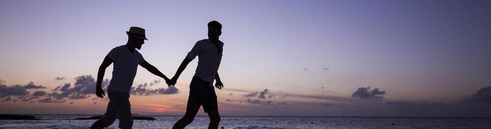 Two people holding hands and walking along a beach at sunset, with the sky displaying shades of purple and orange and the ocean in the background.