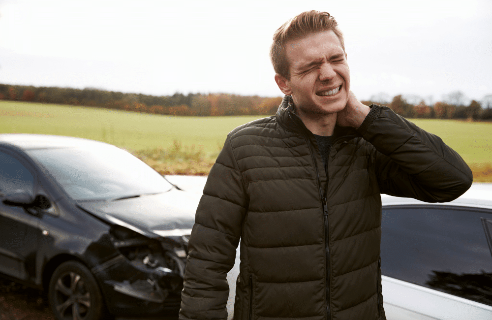 A man stands holding his neck in pain next to two cars that appear to have been in a collision, with visible damage to the front of one car. The scene is outdoors with a field in the background.