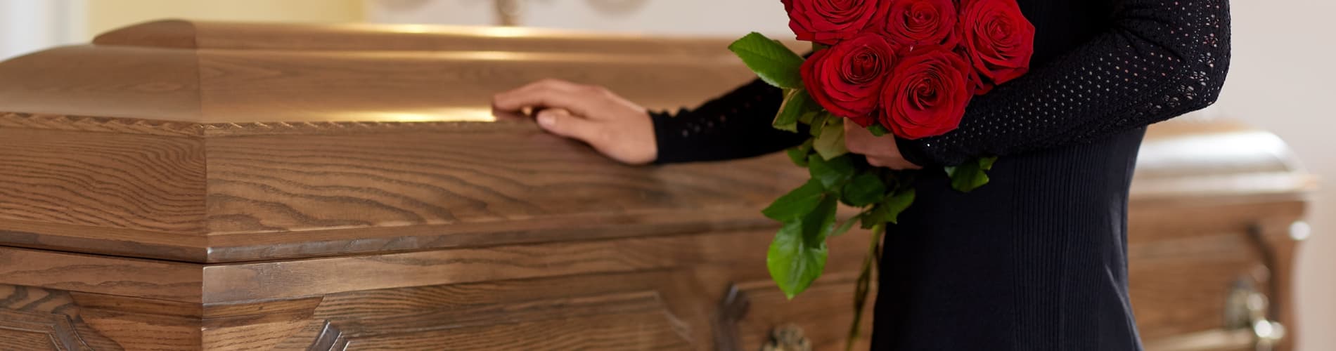 A person in black clothing stands beside a wooden casket, holding a bouquet of red roses, suggesting a funeral or memorial service.