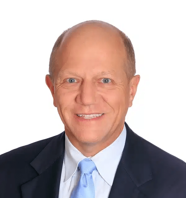 A smiling, middle-aged man with short, light hair wearing a dark suit, light blue shirt, and light blue tie against a plain white background.