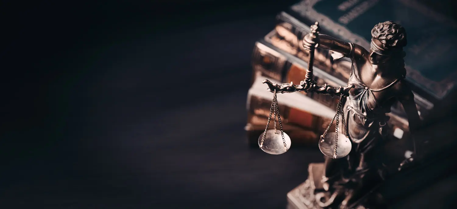 A bronze statue of Lady Justice holding scales stands next to a stack of old law books on a dark surface, symbolizing justice and legal concepts.