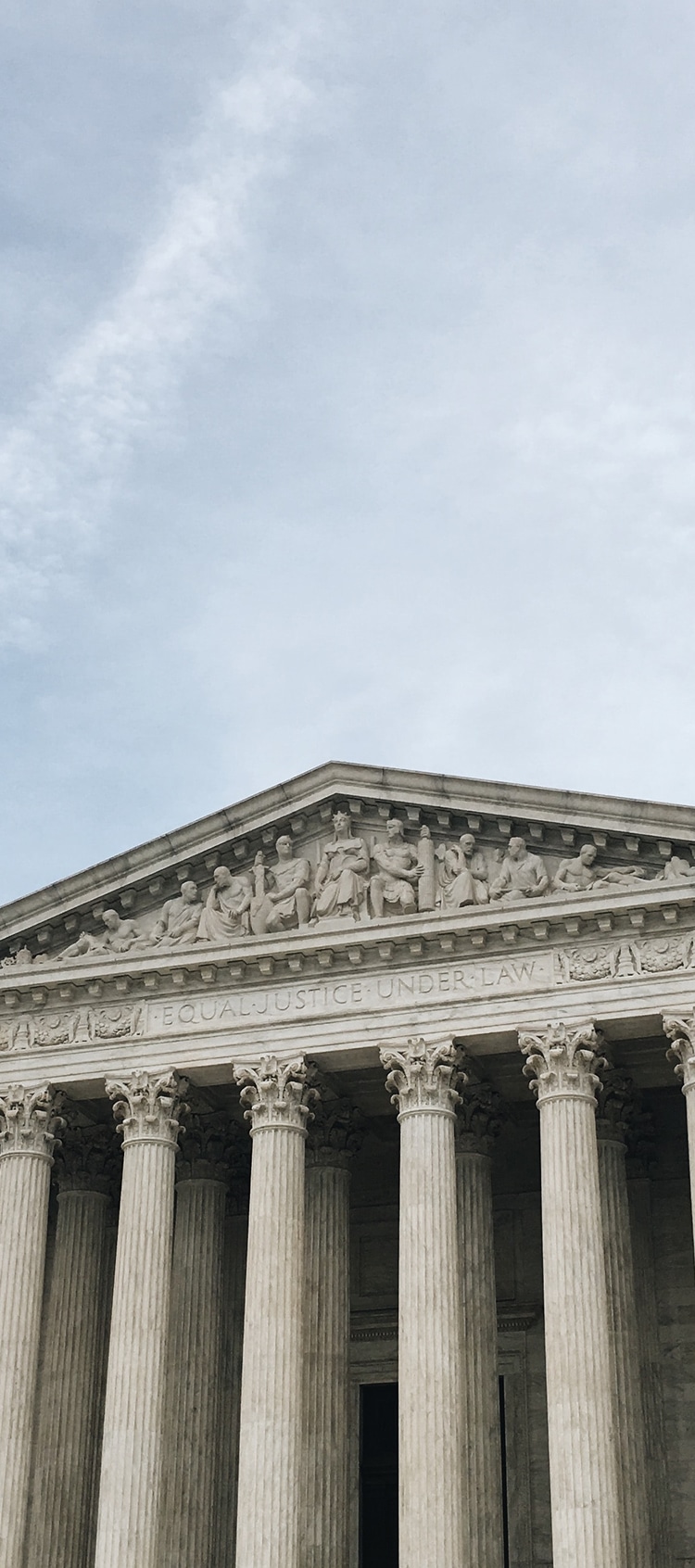 The facade of a classical government building with tall columns and a sculpted pediment. The phrase “EQUAL JUSTICE UNDER LAW” is inscribed above the entrance. The sky is lightly clouded above.