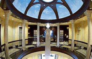 A grand interior with a circular balcony, white columns, and a domed ceiling painted to look like the sky. In the center stands a tall white obelisk, with elegant lighting and decor throughout the space.