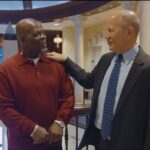 Two men are standing indoors, smiling and talking. One is wearing a red sweater and the other is in a suit and tie, with his hand resting on the others shoulder in a friendly gesture.