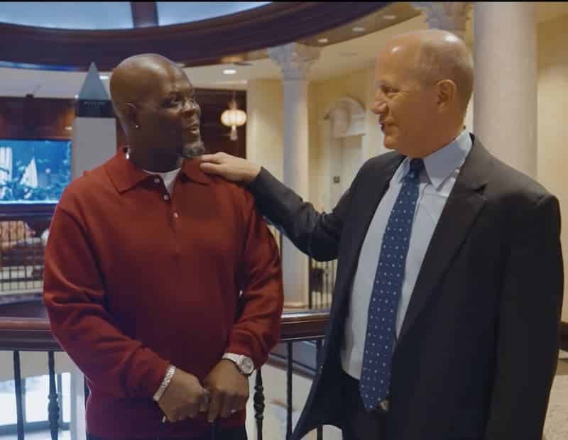 Two men are standing indoors, smiling and talking. One is wearing a red sweater and the other is in a suit and tie, with his hand resting on the others shoulder in a friendly gesture.