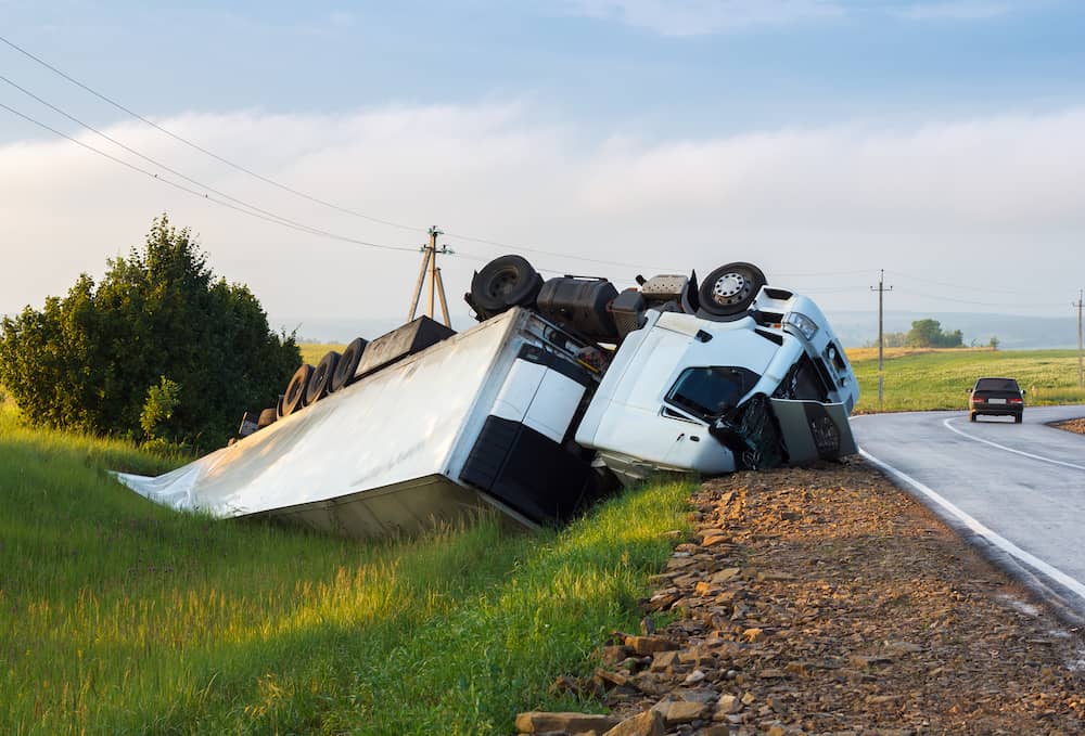 A large white semi-truck lies overturned off a rural road, with its trailer on the grass and cab partially on the roadside. A car drives away in the distance; power lines and fields are visible under a cloudy sky.