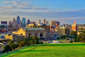A vibrant cityscape of Kansas City, featuring Union Station in the foreground, a fountain, green park areas, and the downtown skyline with tall buildings under a blue, partly cloudy sky.
