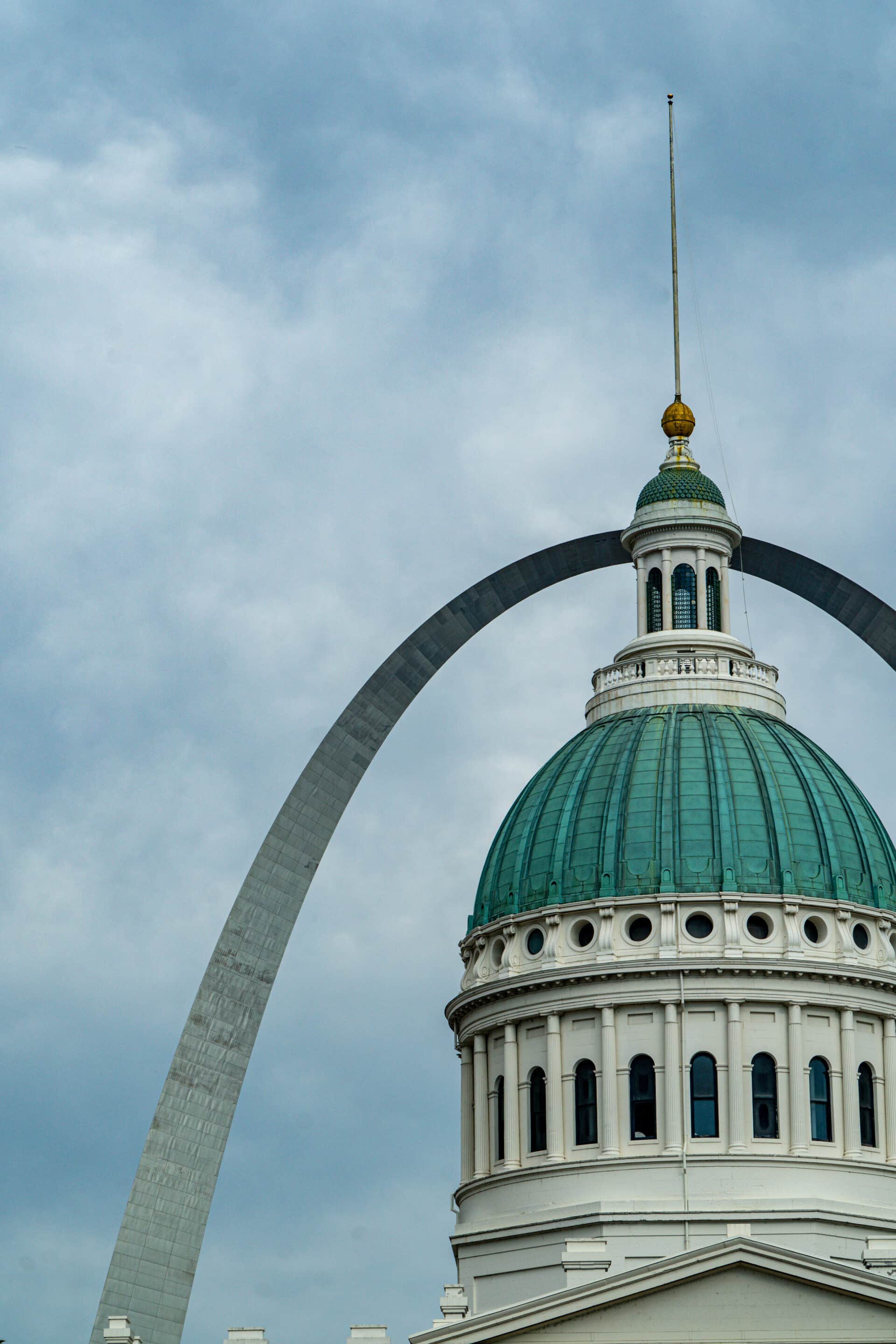 A domed white building with a green roof stands in front of the Gateway Arch in St. Louis, Missouri, under a cloudy sky.