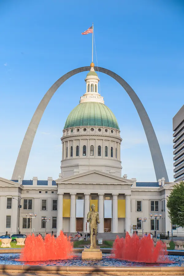 The Old Courthouse in St. Louis with a statue in front, red fountain, and the Gateway Arch rising behind the building under a clear blue sky.