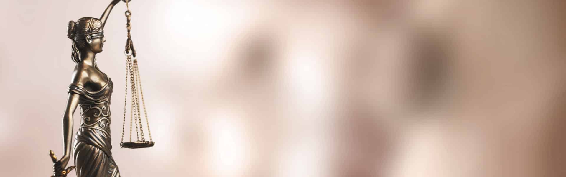 Bronze statue of Lady Justice holding balanced scales and a sword, with a blurred light brown background.