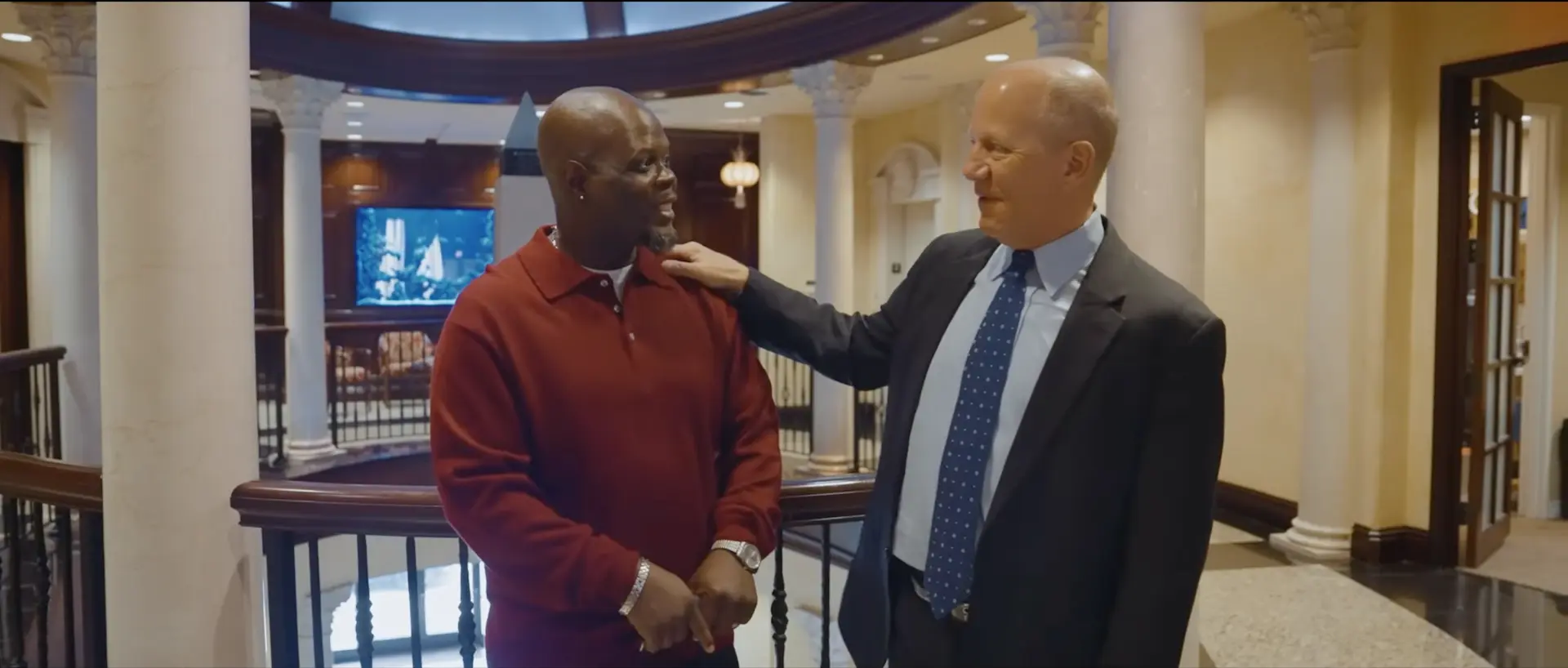 Two men stand indoors, one in a red shirt and the other in a suit and tie. The man in the suit smiles and rests his hand on the other’s shoulder. They appear to be having a friendly conversation in an elegant setting.