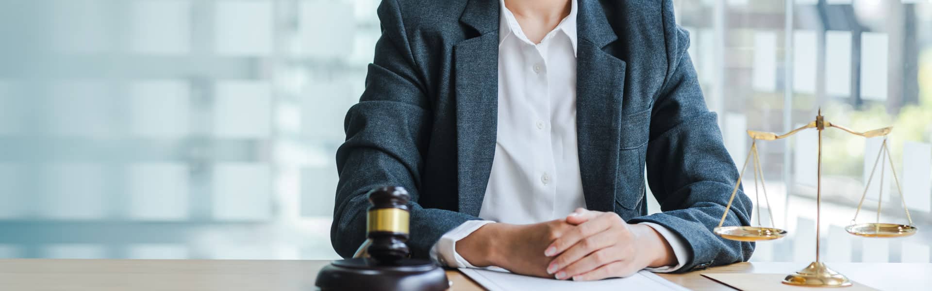 A person in a suit sits at a desk with hands folded on paperwork, next to a gavel and a set of golden balance scales, symbolizing law and justice.