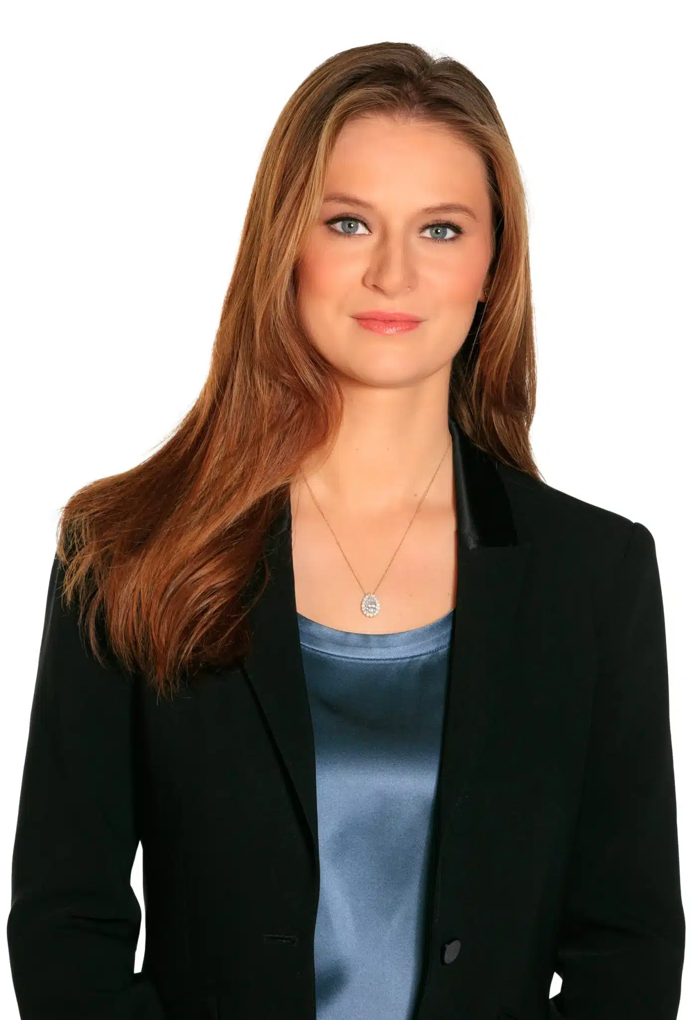 A woman with long light brown hair wearing a black blazer over a blue satin top and a silver pendant necklace, standing against a plain white background.