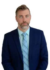 A man with light brown hair and a beard, wearing a navy blue suit, light blue shirt, and patterned blue tie, stands against a plain white background, looking at the camera with a neutral expression.