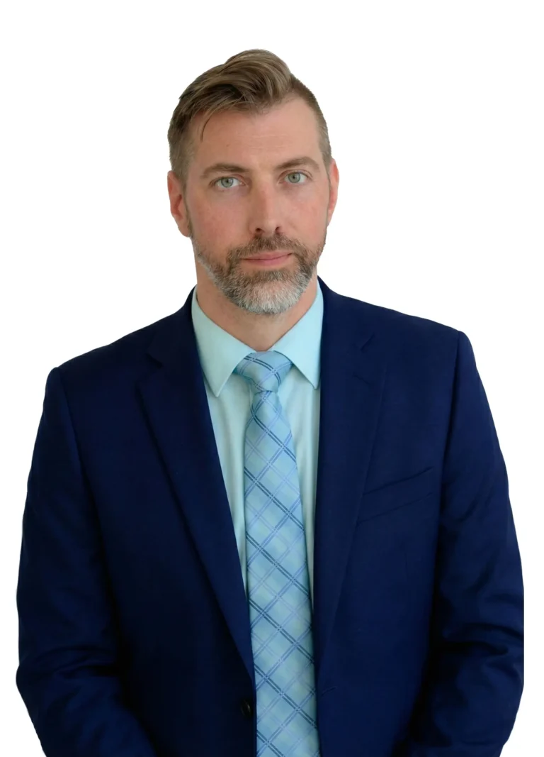 A man with light brown hair and a beard, wearing a navy blue suit, light blue shirt, and patterned blue tie, stands against a plain white background, looking at the camera with a neutral expression.